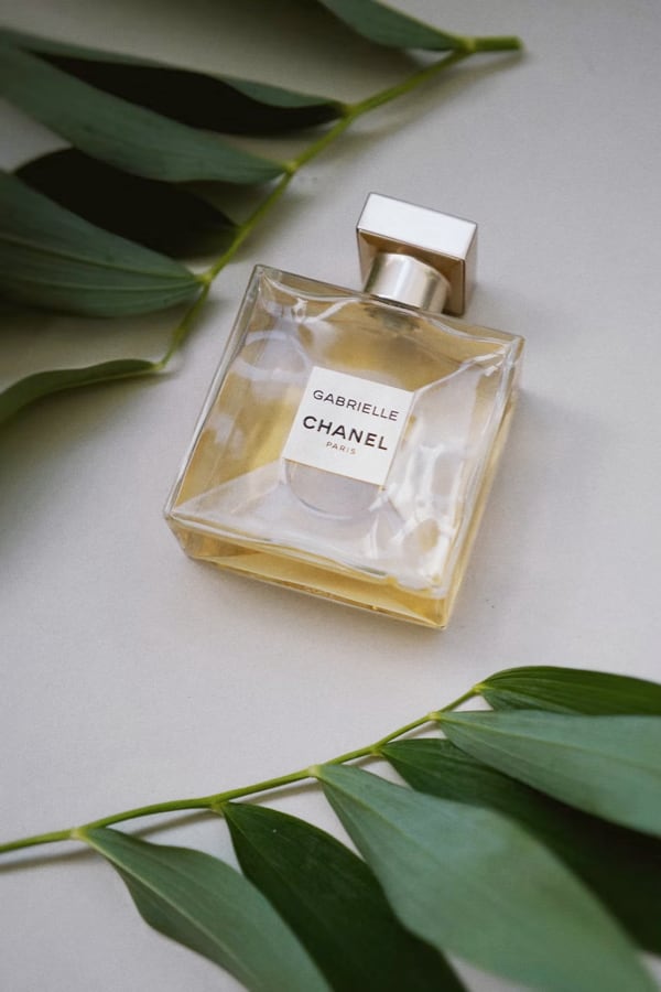 Gabrielle Essense perfume bottle laying flat on a table with green leaves above and below it